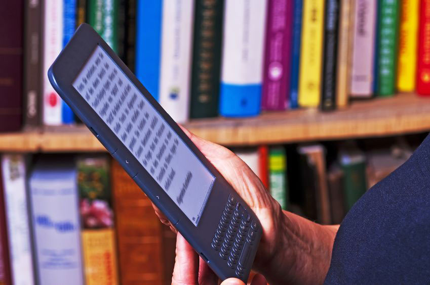 Een persoon houdt een e-reader (ipad) vast. Op de achtergrond een volle boekenkast.