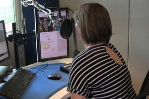 Een voorlezer in een opnamecabine met beeldschermen en microfoon leest een schoolboek voor.