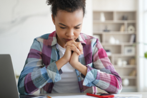 vrouw met ineengevouwen handen onder de kin in gedachten en zittend aan bureau